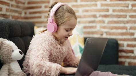 Adorable Blonde Girl Using Laptop And Headphones Sitting On Sofa With Teddy Bear At Home Stock