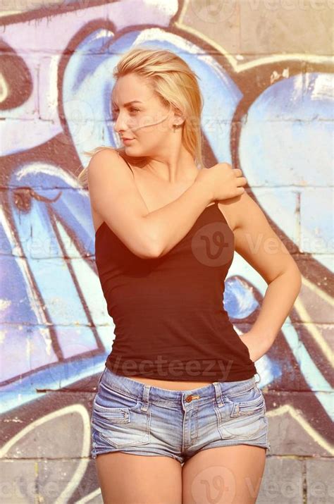 Sexy Caucasian Blonde Girl In Denim Shorts And Black Tank Top Posing Against Graffiti Wall In