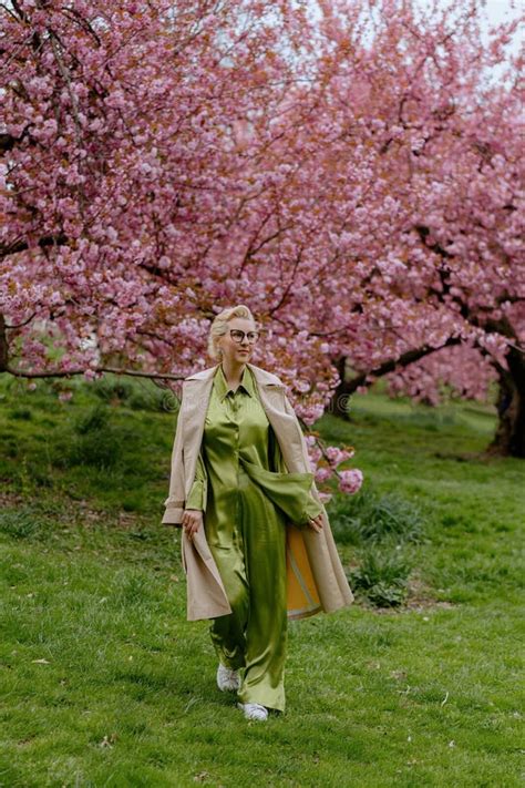 A Blonde Woman In Green Outfit Poses Surrounded By Cherry Blossoms Outdoors Stock Photo Image