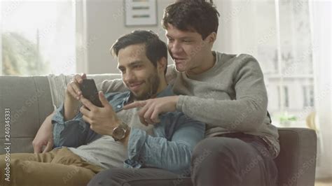 Cute Male Gay Couple Spend Time At Home They Are Lying Down On A Sofa And Use A Smartphone