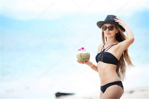 Mujer en bikini en sombrero negro y traje de baño negro bebidas jugo de iluminación de coco en