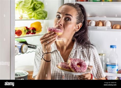 Hungry Brunette In Pajamas Enjoys Sweet Donuts Late At Night By The Open Refrigerator Stock