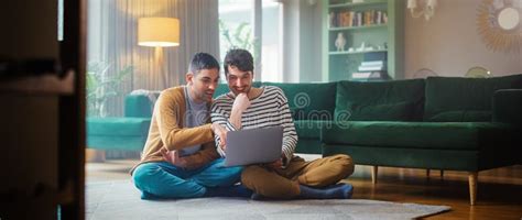 Handsome Gay Couple Using Laptop Computer While Sitting On A Living Room Floor In Cozy Stylish