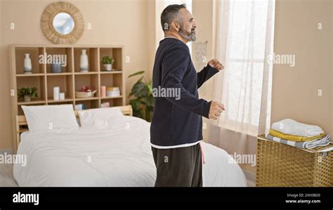 A Mature Man With A Beard And Grey Hair Is Stretching In A Bedroom With A Bed And Bookshelf