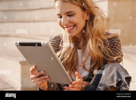 Jeune femme blonde souriante tenant une tablette tout en étant assise sur un escalier dans la