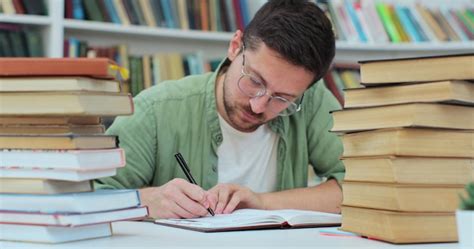 Premium Photo | Man student in library sits at desk full of books Guy wearing glasses preparing ...