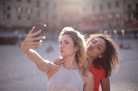 Blonde Woman Behind A Black Haired Lady Taking A Selfie In Front Of The Buildings Stock Photo By