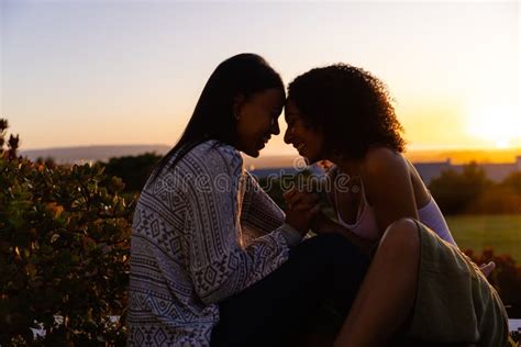 Biracial Lesbian Couple Sitting And Holding Hands In Garden At Sunset Copy Space Stock Image