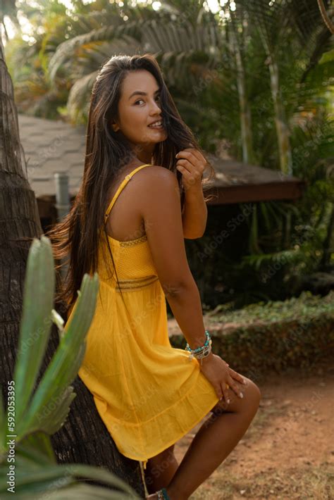 Smiling Flirting Brunette Woman In Yellow Dress Lean On Palm Tree Look Camera And Touch Hair In