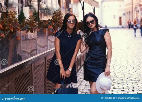 Two Brunette Wearing Stylish Black Dresses In Sunglasses Posing Near A Terrace Cafe In A City