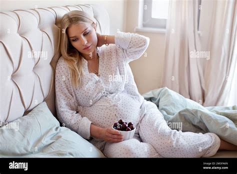 Pregnant Woman Sitting On Bed And Eating Cherry Berries Thoughtful Blonde Model In Pajamas