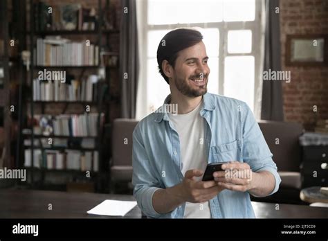 Cheerful Dreamy Millennial Smartphone User Man Holding Cell Stock Photo