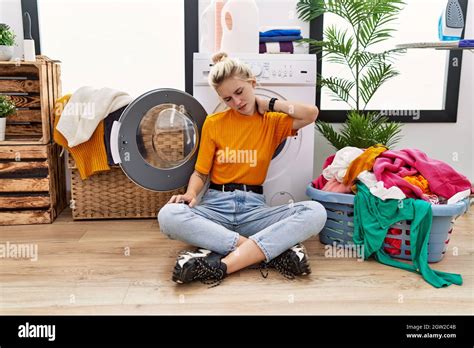 Junge blonde Frau ihre Wäsche wäscht sitzt an der Waschmaschine leidet an Nackenschmerzen