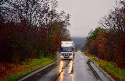 A White Truck Driving Down A Wet Road In The Rain Next To Trees And Bushes