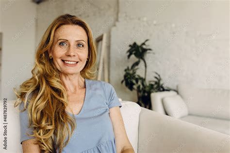 Smiling Mature Woman Sitting On Sofa Against White Home Interior Background Looking Happy