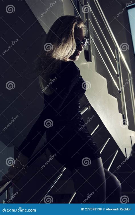 Silhouette Of A Blonde Total Black In A Short Dress Stockings Stands On The Stairs Leaning On