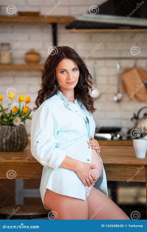 Beautiful Pregnant Brunette Woman In A Shirt Sits In The Kitchen Stock