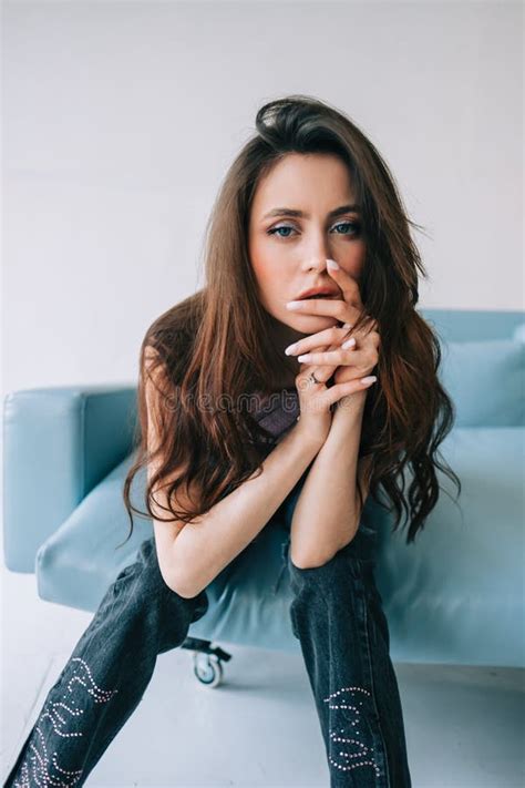 Attractive Stylish Caucasian Woman With Long Brunette Hair Posing On Blue Sofa In Modern Living