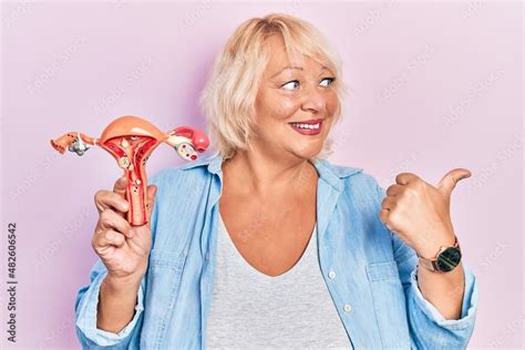 Middle Age Blonde Woman Holding Anatomical Model Of Female Genital Organ Pointing Thumb Up To