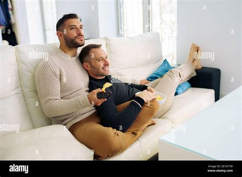 Gay Couple Sitting On The Couch At Home Watching Something On Tv And Having A Snack Stock Photo