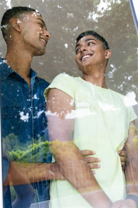 Diversa Pareja Gay Masculino De Pie Por La Ventana Sonriendo Y Abrazando Foto De Archivo