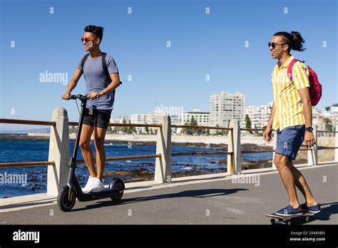 Happy Biracial Gay Male Couple Riding Scooter And Skateboard On Promenade By The Sea Stock Photo