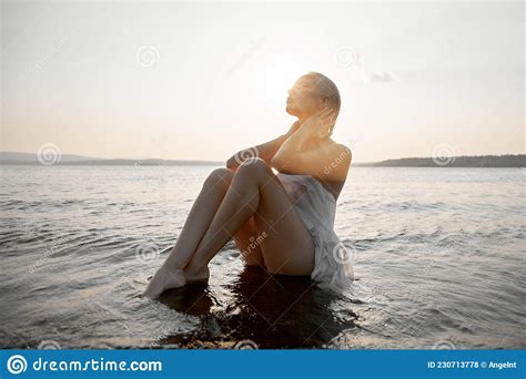 Art Nude Blonde With Short Haircut Is Sitting In Water On Shore Beach Of Lake At Sunset Wet