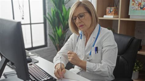 Elderly Female Doctor With Blonde Hair Working At A Computer And Writing In A Medical Office