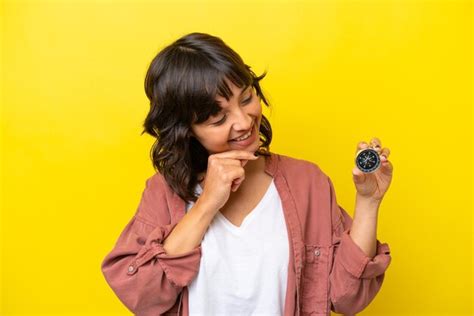 Joven latina sosteniendo una brújula aislada de fondo amarillo mirando hacia un lado y sonriendo