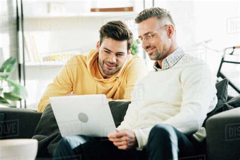 Son Looking At Cheerful Mature Father Using Laptop On Sofa At Home