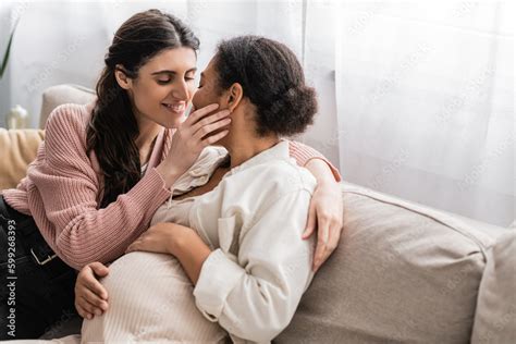 Happy Lesbian Woman Kissing Pregnant Multiracial Partner And Sitting On Couch Stock Photo
