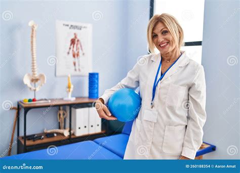 Middle Age Blonde Woman Wearing Physiotherapist Uniform Holding Ball At Physiotherapy Clinic