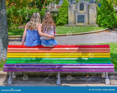 A Couple Of Lesbian Girls Embraced And Sat At A Public Bench Painted With The Rainbow Colors As