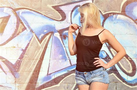 Sexy Caucasian Blonde Girl In Denim Shorts And Black Tank Top Posing Against Graffiti Wall In