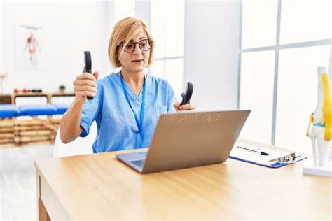 Middle Age Blonde Woman Wearing Physio Therapy Uniform Doing Tele Rehab Using Hand Grip At