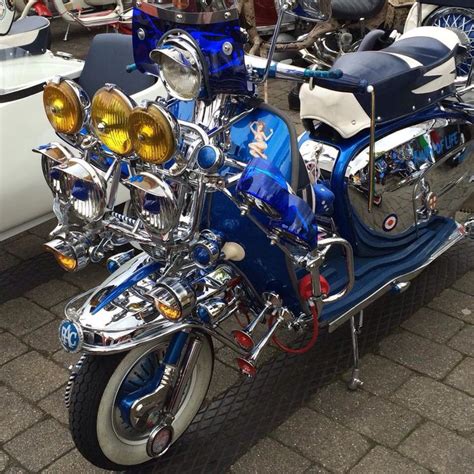 A Blue Motorcycle Parked Next To A White Car On A Brick Road With Other