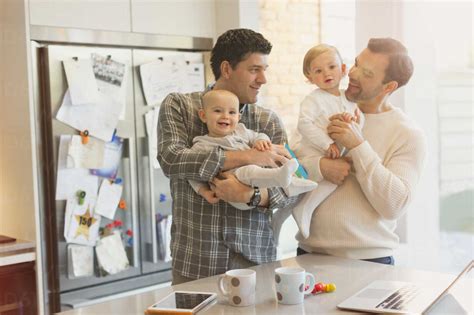 Male Gay Parents Holding Baby Sons In Kitchen Stock Photo