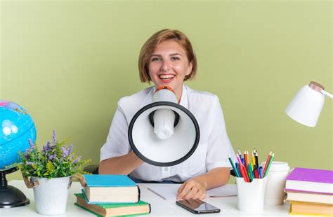 Joyeuse jeune étudiante blonde assise au bureau avec des outils scolaires regardant la caméra