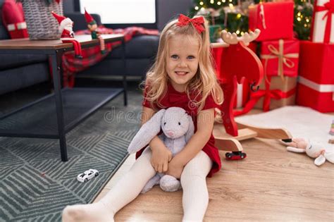 Adorable Blonde Girl Smiling Confident Sitting On Floor By Christmas Tree At Home Stock Image