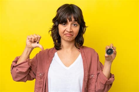 Jovem mulher latina segurando bússola isolada em fundo amarelo mostrando o polegar para baixo