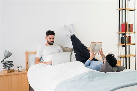 Pareja gay joven tumbada en la cama leyendo el libro y usando el teléfono móvil Foto Gratis