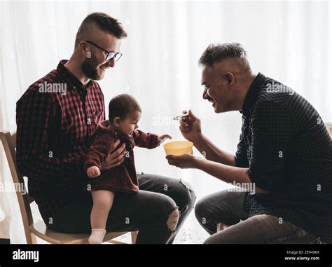 Male Gay Couple With Adopted Baby Girl At Home Two Handsome Dads Feed The Baby Girl On Kitchen