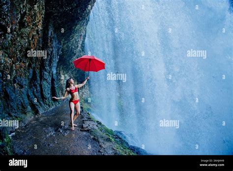 Woman In Red Bikini Holding Umbrella Beneath A Waterfall On Rocky Path