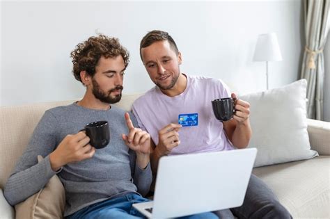Alegre joven pareja gay sonriendo alegremente mientras compra en línea en casa dos jóvenes