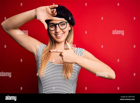 Beautiful Blonde Woman With Blue Eyes Wearing French Beret And Glasses Over Red Background