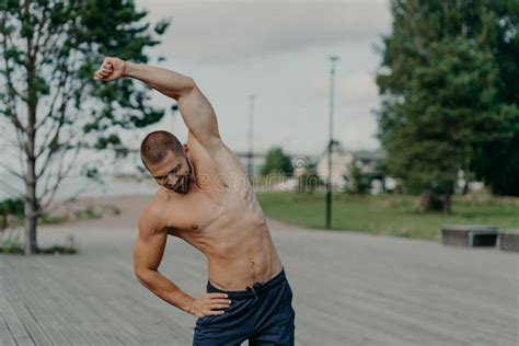 Outdoor Shot Of Active Muscular Man Makes Stretching Excerises And Poses With Naked Torso
