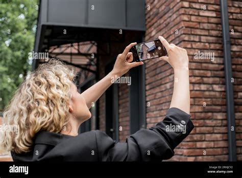 Blonde Real Estate Agent With Mobile Phone Taking Photo Of Modern House On City Street Stock