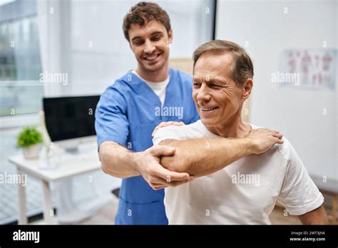Jolly Attractive Doctor Helping His Mature Patient To Stretch During Rehabilitation In Hospital