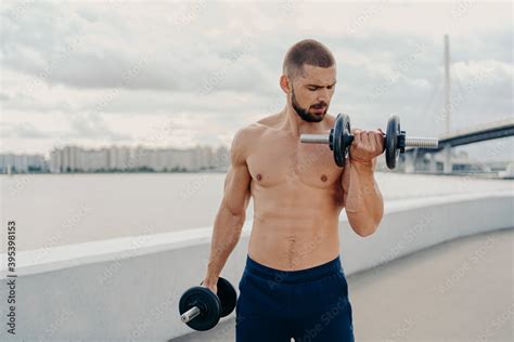 Muscular Guy With Naked Torso Exercises With Barbells Stands Shirtless Trains Muscles Outdoor