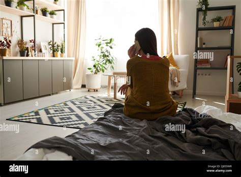 Rear View Of Brunette Woman In Cardigan Sitting On Bed And Calling Close Friend By Cellphone In
